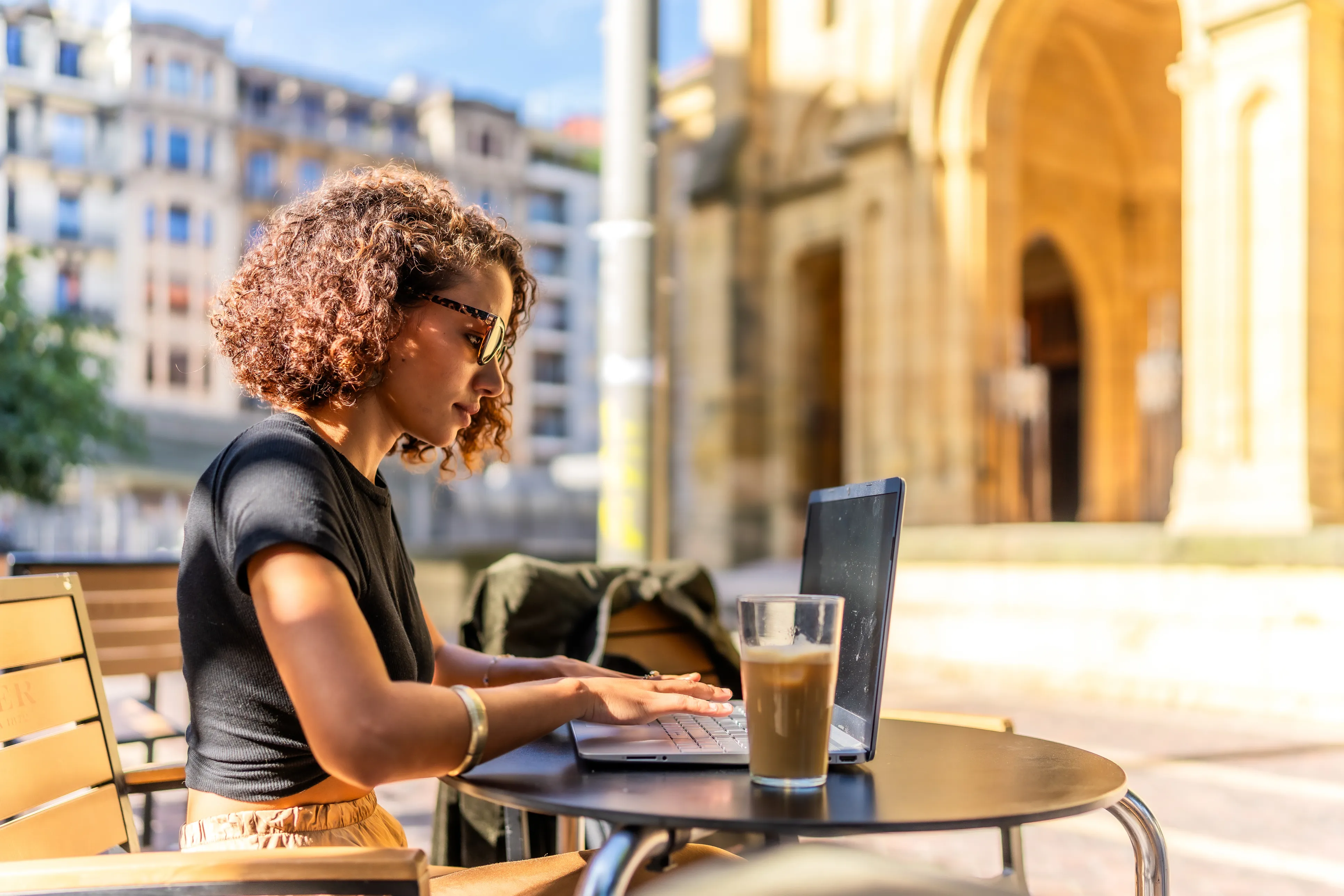 Femme travaillant sur un ordinateur portable à la terrasse d’un café en ville, avec un verre de café posé sur la table.